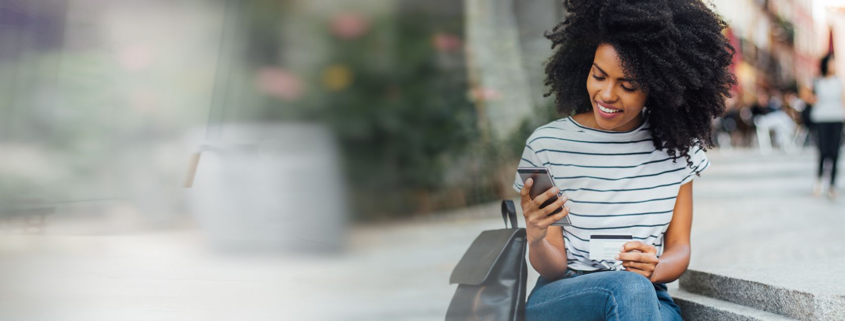 A stylish woman holds her debit card as she logs into Patelco Online on her phone.