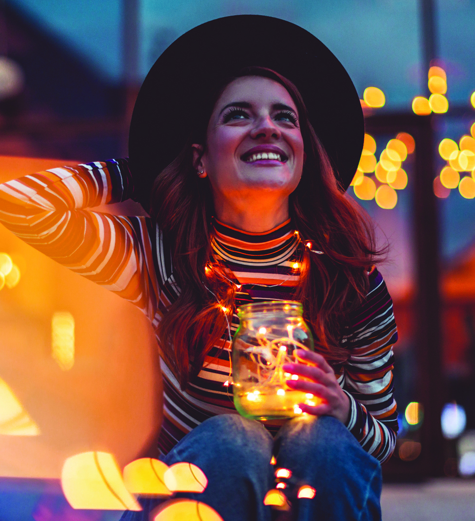 Photograph of person looking up at the sky and smiling with string lights around her